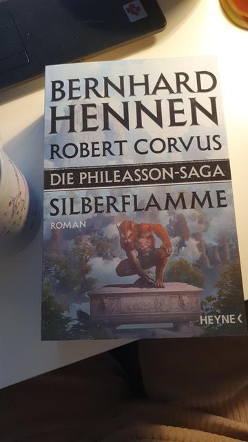 The Book "the Phileasson-Saga" lies on a white table with a coffee mug next to it. 
On the cover is a werewolf in front of a forest, crouching on some kind of altar