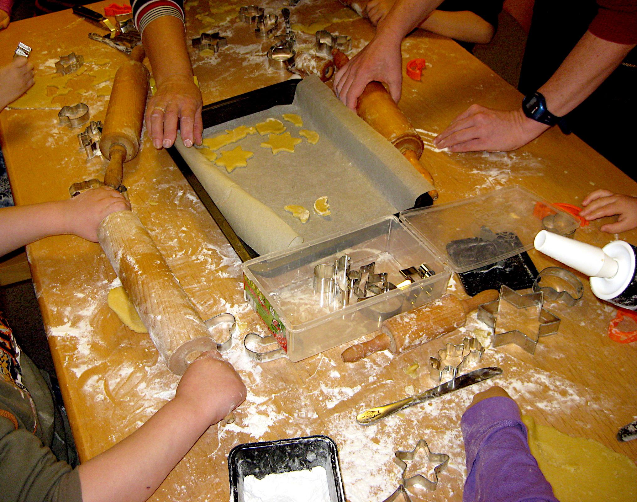 Auf einem Tisch aus Holz steht in der Mitte ein Backblech, auf dem einige ausgestochene Weihnachtsplätzchen liegen. Rund um den Tisch sieht man Arme, die Teig ausrollen oder Plätzchen ausstechen. Es liegen Formen zum Ausstechen herum, der Tisch ist mit Mehl bedeckt.