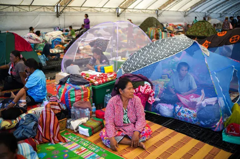 Displaced people take shelter, some inside tents inside the shelter.