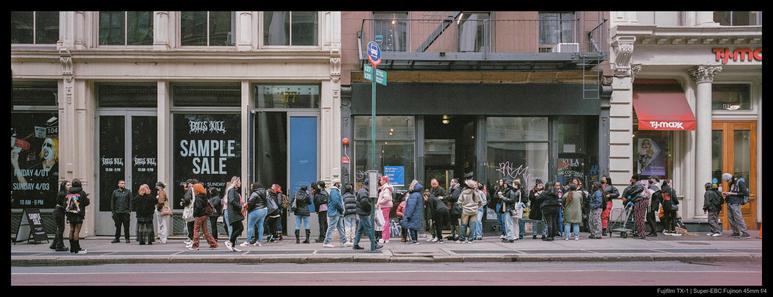 A long line of people gather outside a store preparing to have a sample sale, trailing from the door down the length of another shop and past the edge of the frame.  The crowd is roughly divided in half by a bus stop sign.