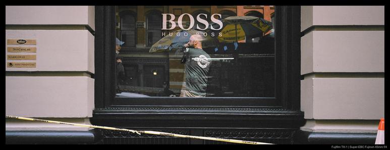 A worker stands behind a storefront window with their hand raised to their mouth as they chat with another worker, barely peeking in from the edge of the windowframe.  The word "BOSS" is emblazoned across the top of the glass over the worker's head.  A strip of caution tape is visible in the foreground.
