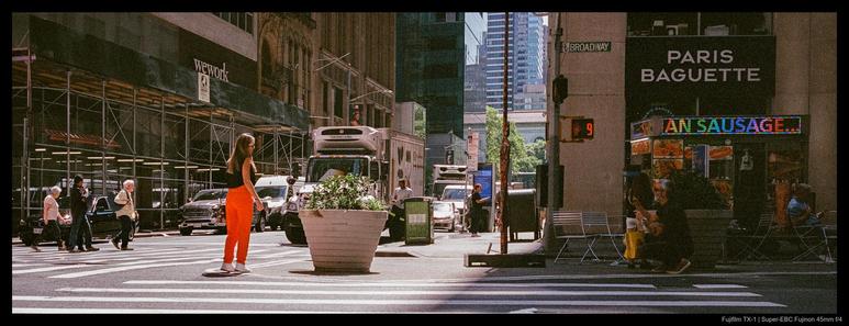 A person dressed in bright orange pants and a black top stands at the edge of a crosswalk near a large planter.  The corresponding signal light shows a nine-second warning light for crossing Broadway.  A Paris Baguette and wework are visible in the background, as is a food cart selling sausages.