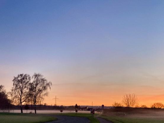 A serene landscape at dawn with a gradient sky transitioning from blue to orange. Silhouettes of trees and a misty field are in the foreground, while a winding road leads into a foggy distance. 