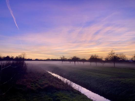 A serene landscape featuring a foggy field at dawn. A calm stream runs through the scene, bordered by trees silhouetted against a vibrant purple and orange sky. Wispy clouds and a faint vapor trail are visible in the sky.