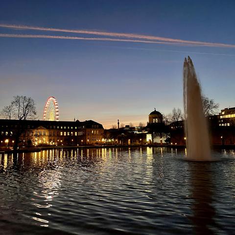 Ein See im Dämmerungslicht, außerdem zu sehen: Kondensstreifen, Gebäude, ua das neue Schloss und das Kunstgebäude, ein Riesenrad und eine Fontäne.
