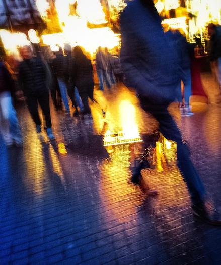 Foto, verwackelt, Abenddämmerung, Weihnachtsmarkt, ein Stand mit vielen gelb leuchtenden Lichtern, im Kontrast dazu vorbeilaufende Menschen in blauem Abendlicht, Bildmitte eine Pfütze, in der sich - verwackelt - das Licht des Standes spiegelt.