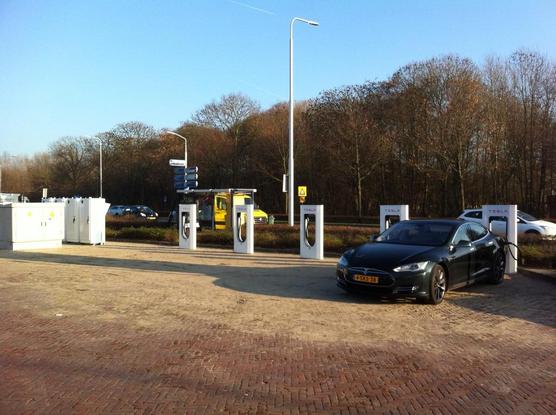 Five white charging dispensers, a dark Tesla Model S next to one of them. On the left side of the image, there is also a transformer box and other technical cabinets.