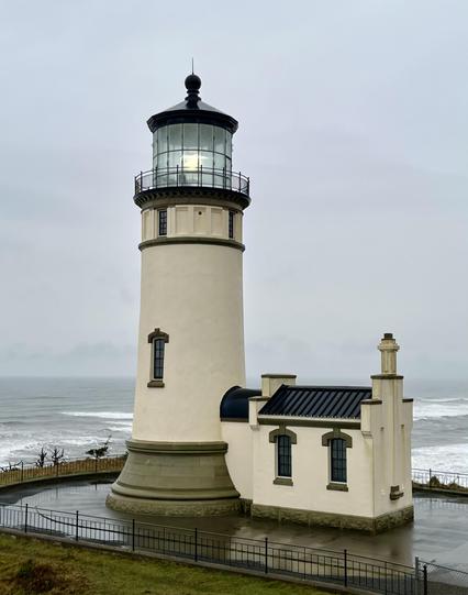 A tall white lighthouse on a bluff above the ocean, a support building at its base and its circling light captured lit up by the camera. White waves can be seen on the
ocean and the concrete footing of the light is wet with rain.