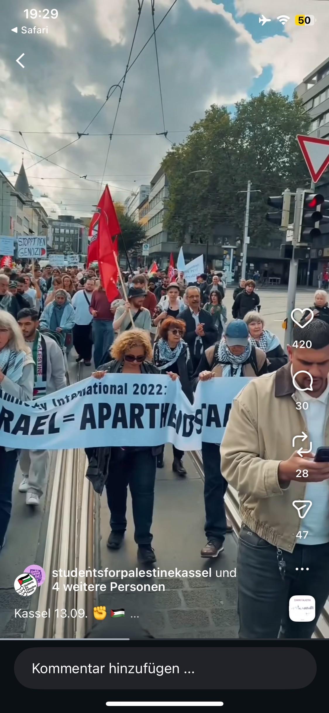 Eine Gruppe von Demonstranten marschiert in einer städtischen Umgebung und hält Banner und Flaggen  mit der Aufschrift "Israel = Apartheidstaat". Die Menge umfasst Personen, die Keffiyehs tragen und Schilder tragen, die ihre Solidarität mit Palästina zum Ausdruck bringen.