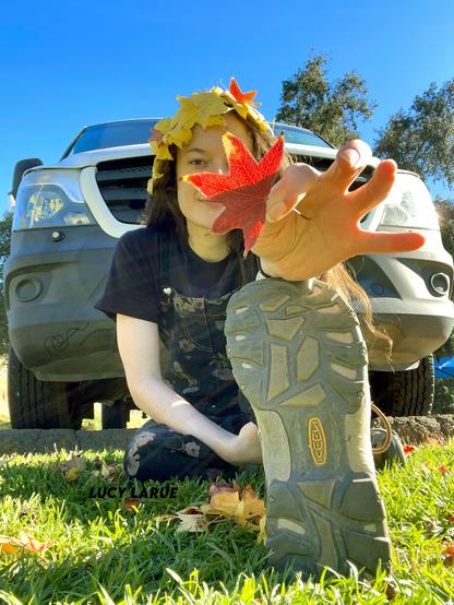 A blue sky sunny day, brunette Lucy LaRue in dark floral overalls with a yellow crown of fall leaves in her hair holds a red leaf out to you, its fronds covering half her smiling face as she looks at you while sitting on the grass in front of her white sprinter van, her leg extended toward you, the sole of her Keen brand hiking boots facing the camera, supporting her leaf arm.