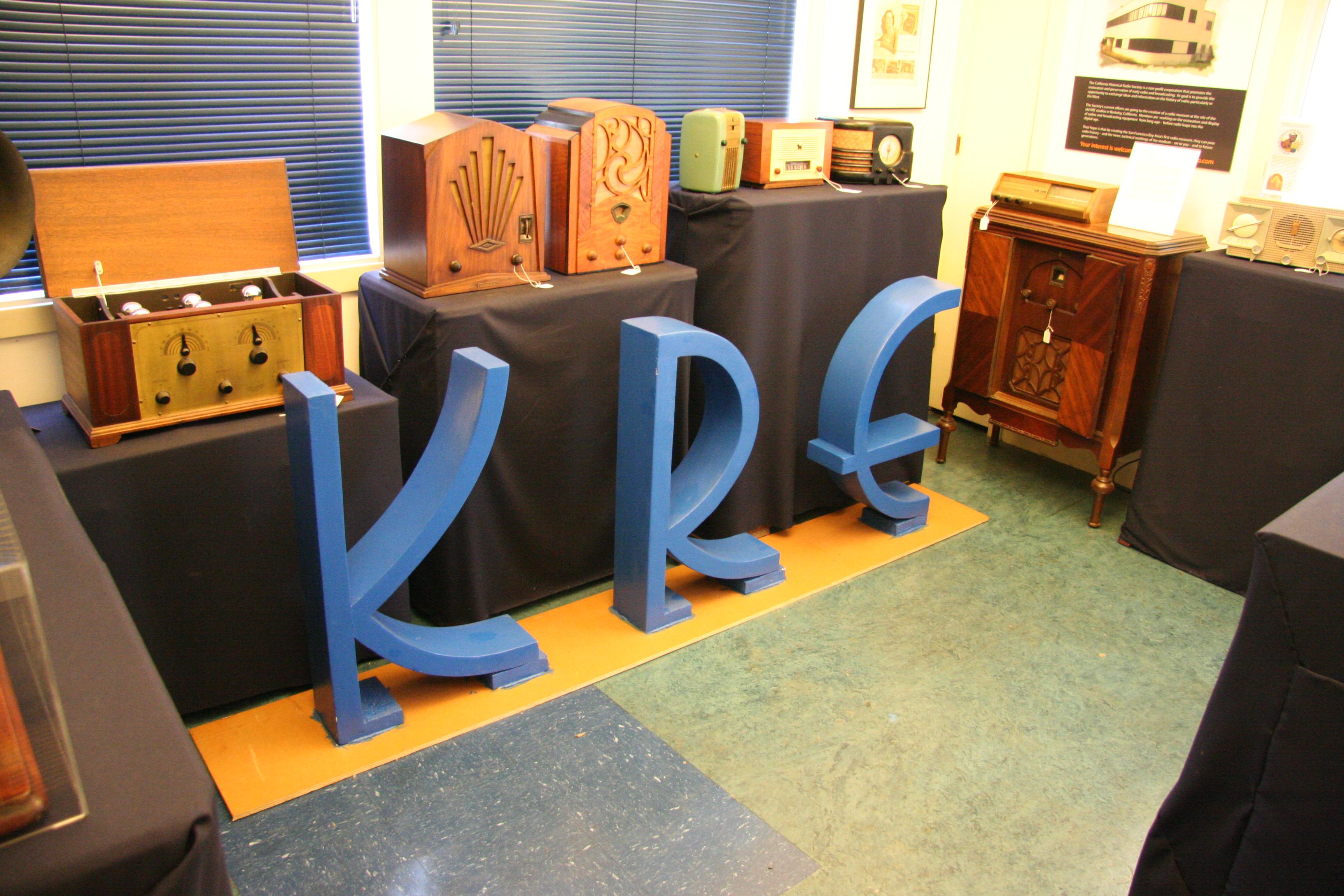 In a small office with a linoleum floor, antique radios are displayed on top of cabinetry, cloth-covered tables, and other radios. A line of cabinets runs parallel to an exterior wall, which has two windows obscured by metal Venetian blinds, and in front of that row of cabinets, sitting on the floor, are the three letters K R E in a very 1950s-looking typeface, each about two and a half feet high. The letters appear to be recently painted blue and mounted to a board.