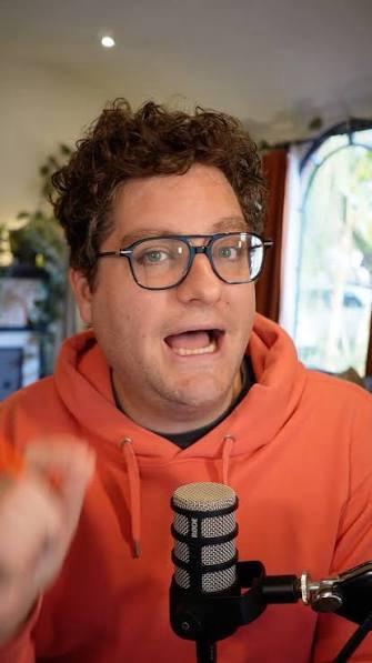 White nerd Alex Falcone wearing glasses, curly hair, orange hoodie with a microphone
