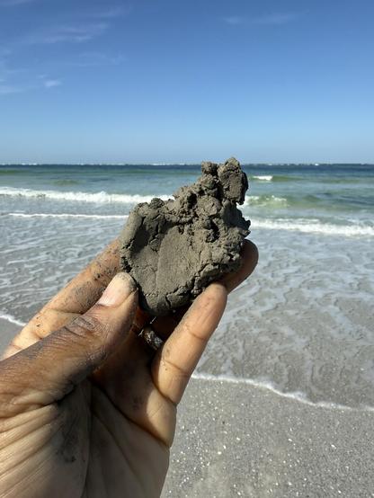 A photo of my hand holding a small amount of moist gray clay with. The blue-green waters of the gulf and a blue sky in the background. I touched it to my tongue and yes, it’s salty. I have another clay sample that I found on the beach in Cape Cod and when I fired it, it formed a glossy coating because salt is a flux that can form a glaze. I wonder if this will do the same thing?