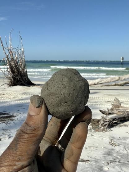 I mixed a bit of gulf water into the clay to moisten it a bit further and formed a ball. The photo is of my hand holding a small imperfect sphere of gray clay. The residue is all over my hands. In the background are white sand, drift wood and palm fronds and the gulf waters as well as a blue sky. 