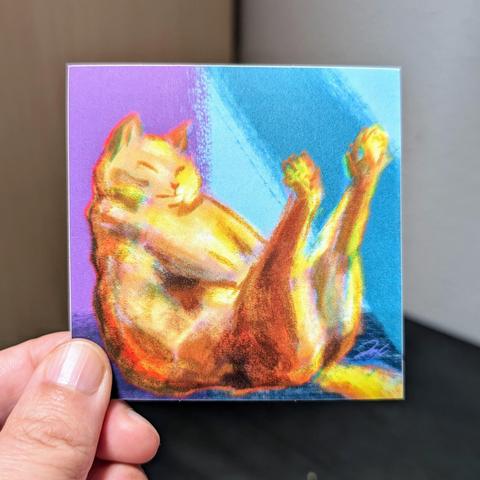 Photo of a left hand holding a square sticker. The sticker is a matte painting of an orange cat in boat pose.