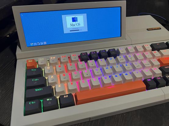 A small computer in beige plastic that resembles a slabtop from the mid 1980s. A rainbow backlit keyboard with beige, dark gray, and orange keycaps is in the front with a small LCD screen on the upper left tilted up toward the viewer. On the screen is the MacOS boot screen with icons of control panels and extensions loading.