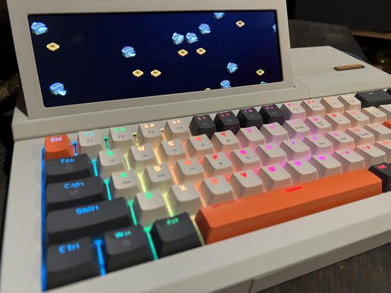 A small computer in beige plastic that resembles a slabtop from the mid 1980s. A rainbow backlit keyboard with beige, dark gray, and orange keycaps is in the front with a small LCD screen on the upper left tilted up toward the viewer. On the screen is flying toasters screen saver from After Dark.