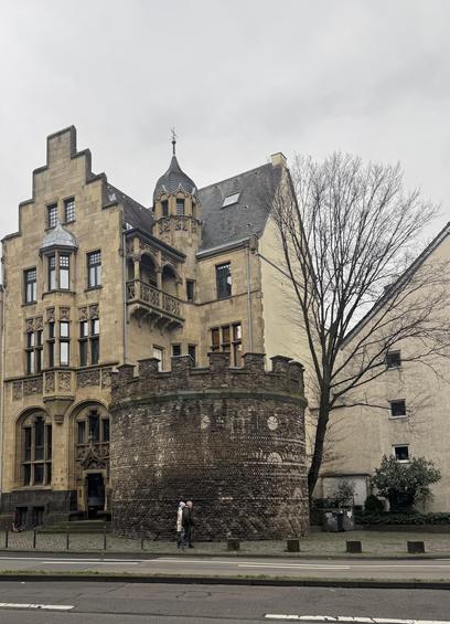 A historic cream and red stone building in Cologne with tall arched windows and decorative details. Two autumn trees stand in front, partly covering the facade in warm sunlight.