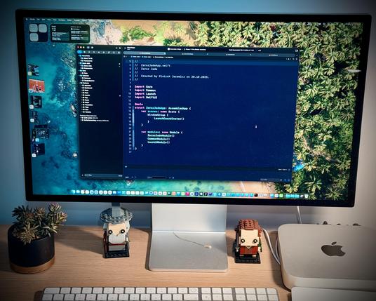 Large monitor showing Swift code editor with macOS dock, desk with Mac mini, keyboard, two Lego minifigure builds and a small potted succulent.