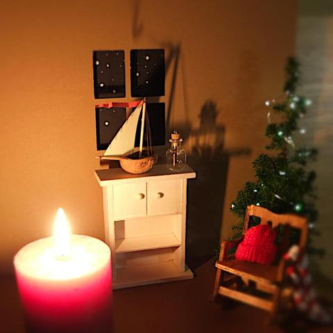 Foto einer Miniaturszene eines weihnachtlich eingerichteten Zimmers, ähnlich einem Puppenhaus. Rechts im Bild ist ein Schaukelstuhl mit Wollmütze und Schal darüber gelegt zu sehen. Dahinter steht ein kleiner Weihnachtsbaum mit Lichterkette. Links unten im Bild ist eine rote lebensgroße brennende Kerze. An der Rückwand ist ein Fenster reingeschnitten mit Sternenhimmelmotiv. Davor steht ein Holzschränkchen mit eine Mini-Glasflasche darauf, nebem einem Modellsegelboot aus einer Walnussschale.