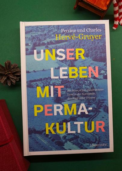 Draufsicht eines Buches mit dem Titel " Unser Leben mit Permakultur". Ringsrum liegt weihnachtliche Dokoration, z.B. ein Tannenzapfen links im Bild, darunter ein dunkelrotes Geschenk.
