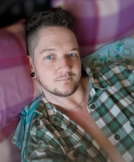 A trans man with brown hair and facial hair. He looks into the camera with piecing blue eyes and a slight smile that is also shown in his eyes, giving a a sense of someone who loves a laugh and is good fun. He wears a green, grey and white plaid shirt, open down the front. He looks comfortable and relaxed as he reclines against pink plaid cushions.