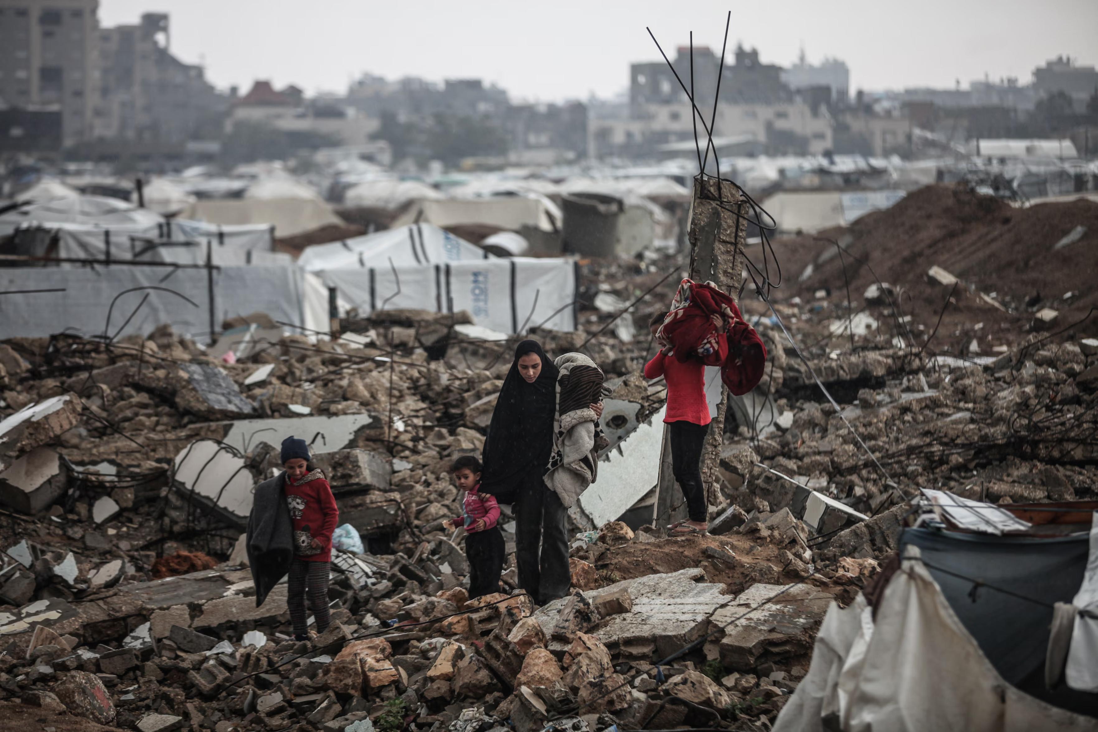 A family with children walk through the rubble behind a tent camp.