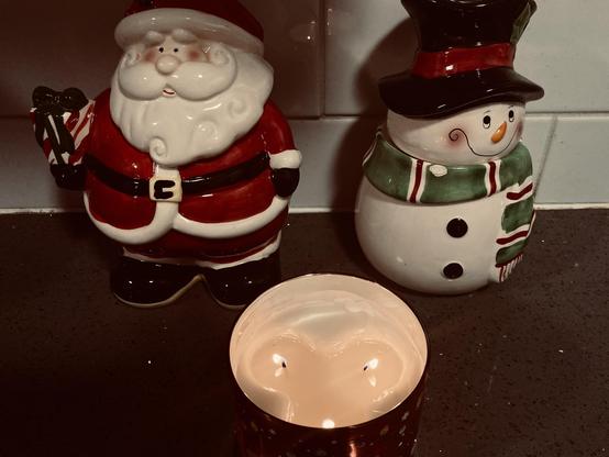 A metallic three wick lit candle on a kitchen counter in front of festive Santa and snowman cookie jars.