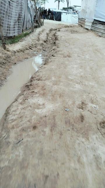 Muddy dirt road with a trench of standing water, flanked by makeshift shelters and tents. A group of people stand in the background.