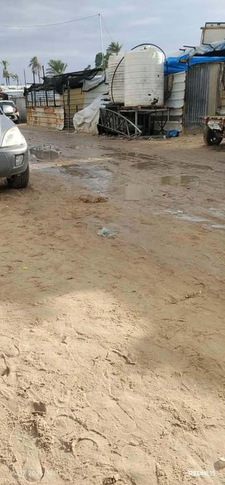 Sandy, wet ground with puddles and a building complex to the right, composed of corrugated metal and white tanks; a partial car frame on the left and a small cart to the right, under a cloudy sky.