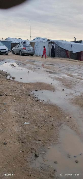 A muddy dirt road with puddles and a row of temporary shelters. A child in a red tracksuit is walking on the road, with two other people standing nearby. Two parked cars are visible. The sky is overcast.