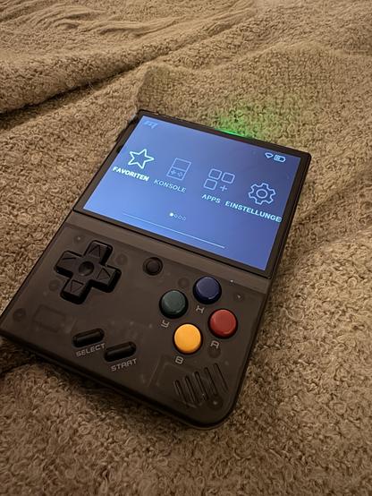 Ein Handheld-Gaming-Gerät wird auf einer strukturierten, hellen Decke angezeigt. Der Bildschirm zeigt eine Benutzeroberfläche mit Optionen, die auf Deutsch gekennzeichnet sind, einschließlich Favoriten, Konsole, Apps und Einstellungen. Das Gerät hat ein Richtungspad, rote, gelbe, grüne Tasten