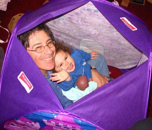 Photograph of Adrian Segar holding his granddaughter in an indoor playhouse. She is waving at the camera.