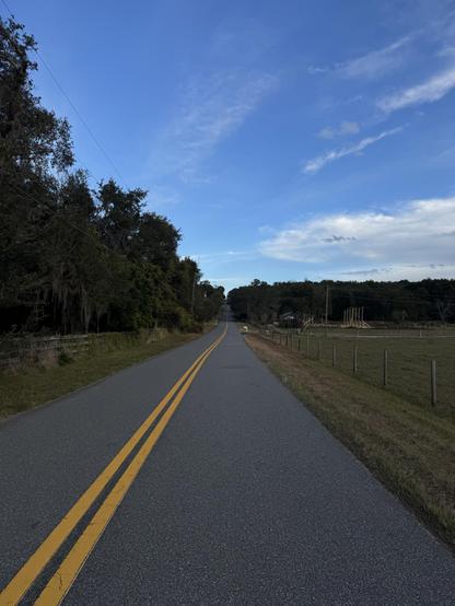 A road with a double yellow line rising up to tree line and blue sky with wispy clouds.