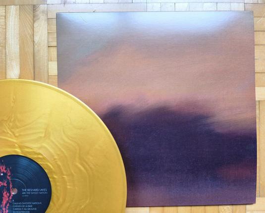 Ein Bild einer goldenen Schallplatte neben ihrem Albumcover, das einen Farbverlauf von warmen Farben von tiefem Violett zu Braun zeigt. Die Schallplatte trägt den Titel „The Besnard Lakes Are the Ghost Nation“. Der Hintergrund besteht aus Holzplanken.
