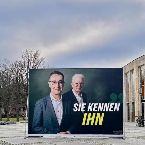 Großplakat mit Fotos von Cem Özdemir und Winfried Kretschmann, links vom Plakat Bäume, rechts das Forum Ludwigsburg, oben Winterhimmel