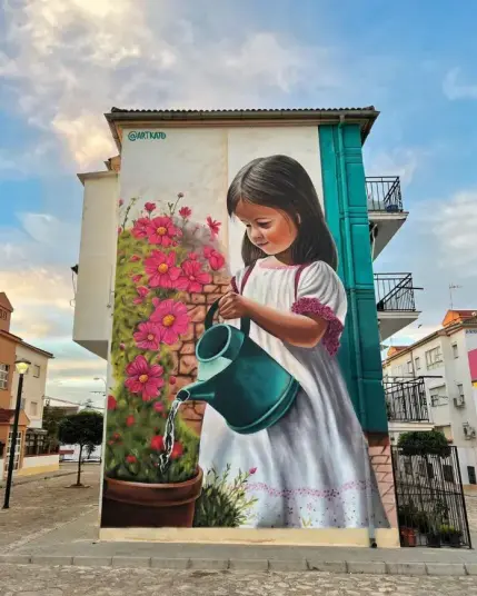 A girl with shoulder-length black hair wearing a dress. She has a watering can in her hand and is using it to water the flowers. The flowers are red.Street art on the wall of an apartment building.
