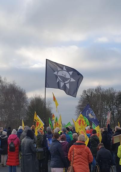 Demonstrierende auf der Straße vor dem Atommüll-Zwischenlager Ahaus, darunter u.a. "Atomkaft? Nein, danke!"-Fahnen und eine schwarze Fahne mit einer Anti-Atom-Sonne.