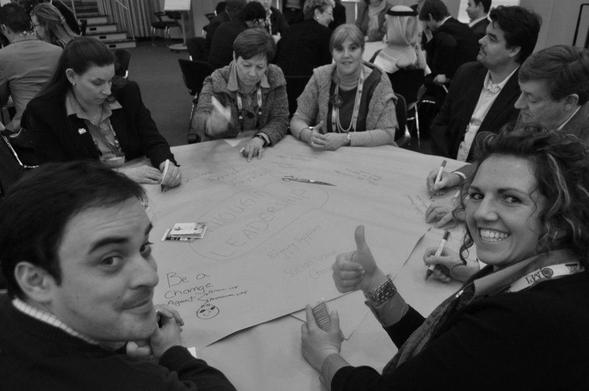 A black and white photograph of participants seated at a round table during a Solution Room session, with one of them giving the thumbs-up sign
