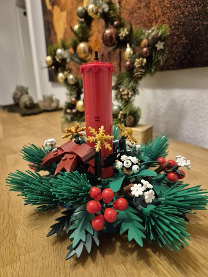 This image features a festive holiday centerpiece made from building blocks, designed to resemble a traditional Christmas decoration. At the center of the arrangement is a red candle, surrounded by green pine branches, red berries, white flowers, and gold snowflakes—all constructed from colorful blocks.

The centerpiece is placed on a wooden surface, with a decorated Christmas tree in the background, adding to the holiday ambiance. The tree is adorned with ornaments and garlands, enhancing the …