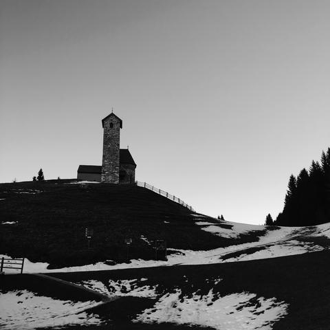 Il prato di montagna mezzo innevato, con una chiesetta ed il suo alto campanile posizionata sulla cima della collina al centro. In cielo non si vedono nuvole