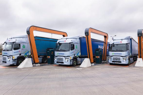 Three electric trucks are parked at a charging station, with modern charging infrastructure and a cloudy sky in the background. The trucks display branding for a company, emphasizing their electric nature.