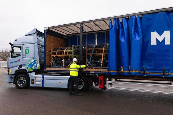 A truck is parked with its blue tarpaulin curtain partially opened, revealing wooden pallets holding machinery or equipment. A worker in a yellow safety vest and hard hat is adjusting straps inside the truck. The scene is set on a wet road under a cloudy