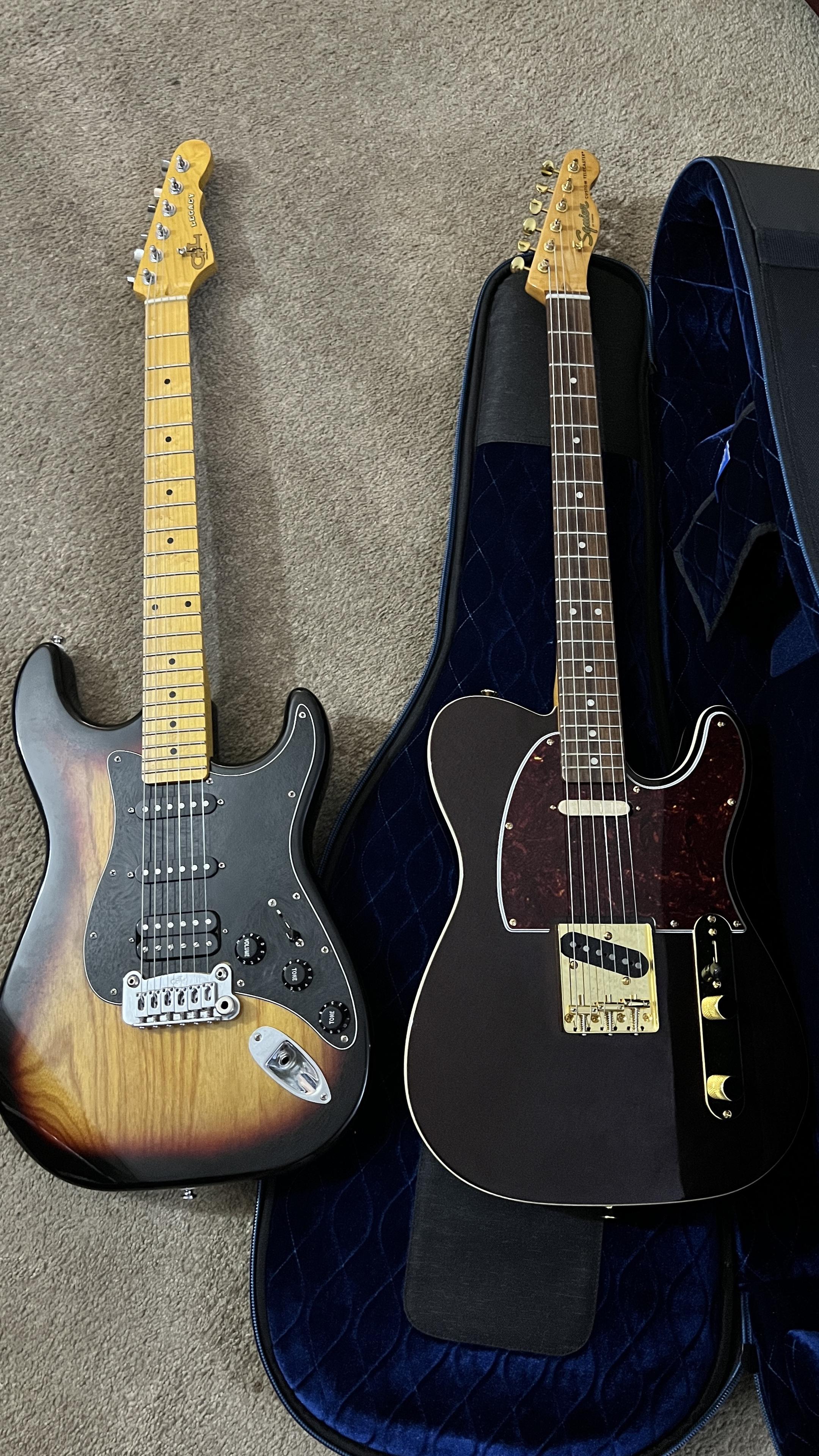 Two fender squire electric guitars are displayed on a carpet: a sunburst electric guitar with a solid body on the left and a black telecaster-style guitar on the right, partially inside a padded guitar case.