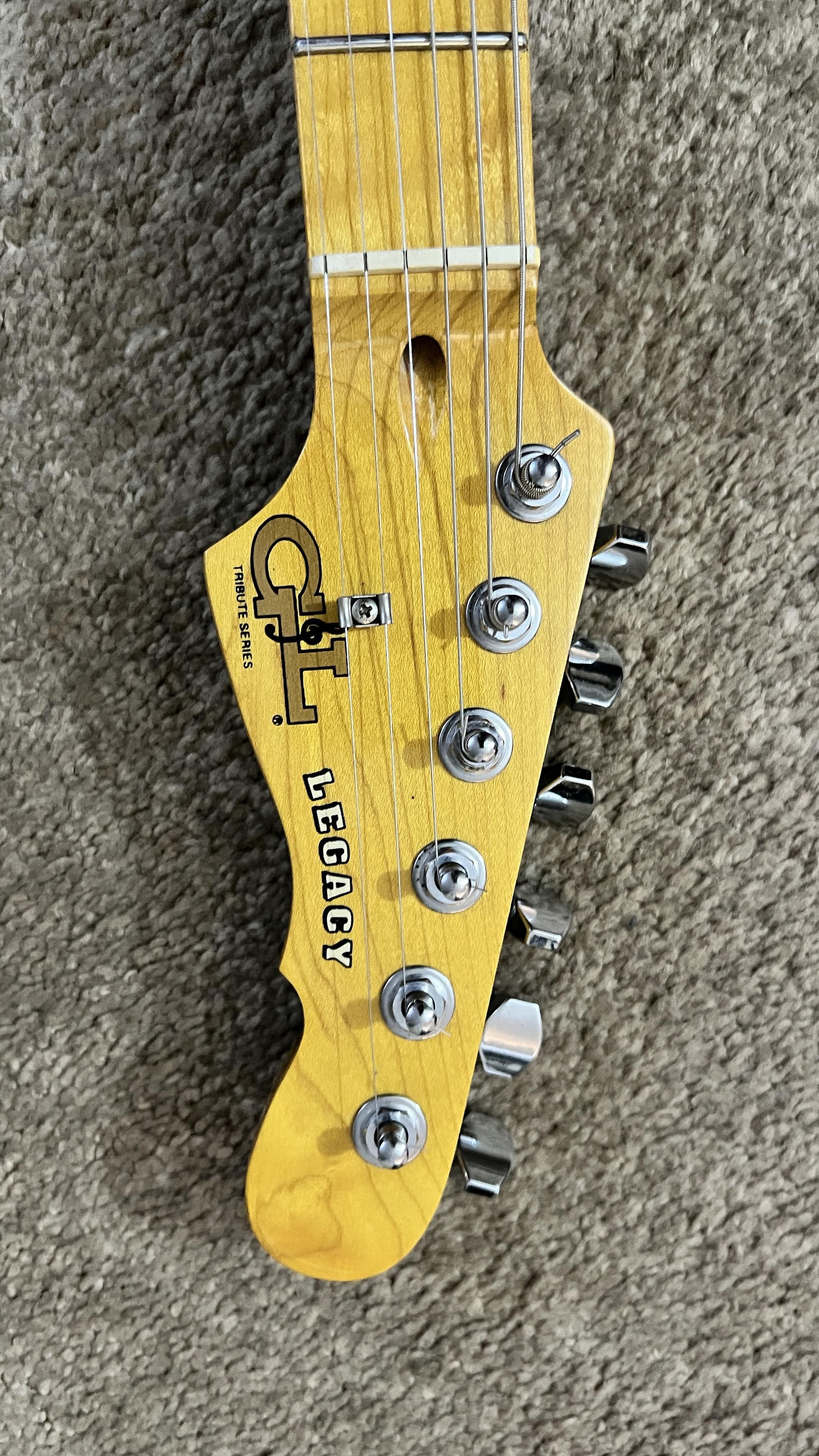 The image shows the headstock of a fender electric guitar featuring a natural wood finish, metal tuning pegs, and branding that reads "G&L TRIBUTE SERIES LEGACY." The background is a textured brownish gray surface.