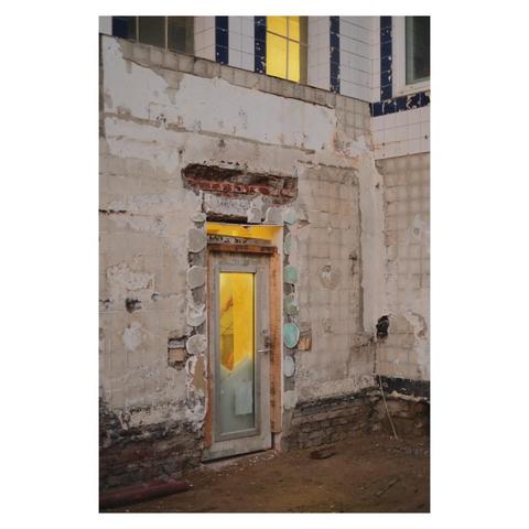 Foto einer Hausfassade während der Renovierung. Der Putz ist abgeschlagen und vereinzelt sind Reste und Spuren von alten Fliesen zu sehen. Eine Glastür durch die warmes, gelbes Licht scheint ist Provisorisch eingebaut. Der gelbe Lichtschein wiederholt sich in einem Fenster im ersten Stock. Zwei weiter Fenster dagegen sind dunkel. 