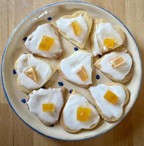 Auf einem Teller aus Ton und mit blauen Punkten sowie blauem Rand liegen herzförmige Plätzchen mit weißer Glasur sowie kandierter Orange (eins kann auch Orangeat dazu sagen) darauf. Sie sind kreisförmig angeordnet und in der Mitte liegt noch eines.