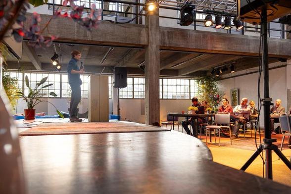 Person in a distance standing on stage with a microfone. Some audience. The setting is a old manufacturing hall