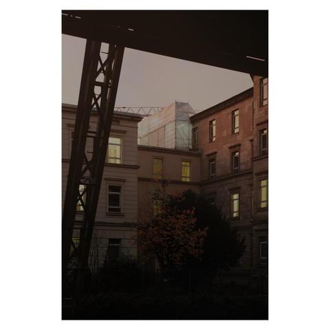 Foto einer urbanen Szene bei Dämmerung: Im Vordergrund befindet sich ein dunkler Metallträger, der diagonal von links oben ins Bild ragt. Dahinter sind mehrere mehrstöckige Gebäude mit hell erleuchteten Fenstern zu sehen. Ein Gebäude ist teilweise mit einem Baugerüst und einer Schutzfolie versehen. Im Innenhof steht ein Baum mit herbstlicher Färbung. Die Atmosphäre wirkt ruhig und leicht melancholisch durch die warmen Lichtakzente und die dunklen Schatten. 