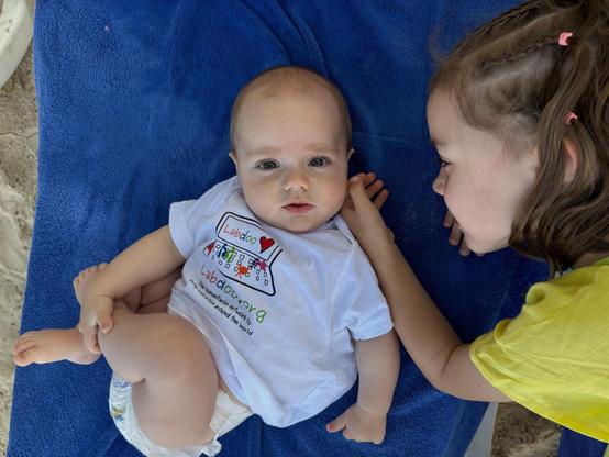 Auf einer blauen Decke liegt ein Kleinkind mit einem T-Shirt von Labdoo. Die größere Schwester liegt rechts daneben und streichelt das Kleinkind. Auf ausdrücklichen Wunsch der Eltern wird das Foto unverpixelt veröffentlicht.